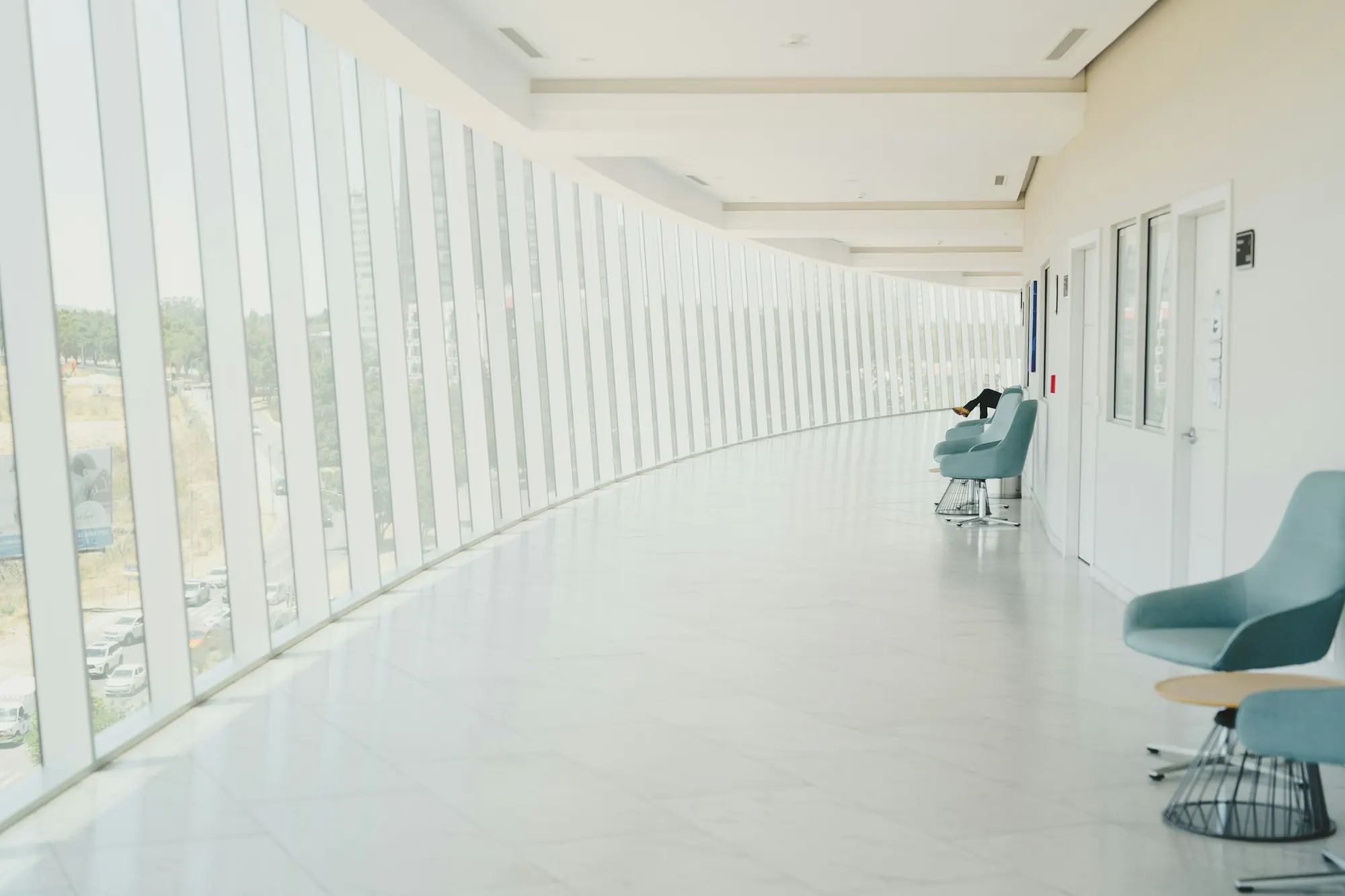 A long, bright hospital corridor curving along a wall of tall windows, with a few blue chairs by the doors